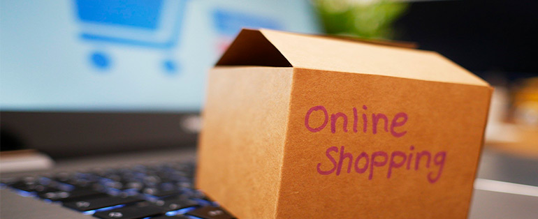 Zoom em caixa de papelão com os dizeres "online shopping" apoiada em um notebook com a imagem de um carrinho na tela.